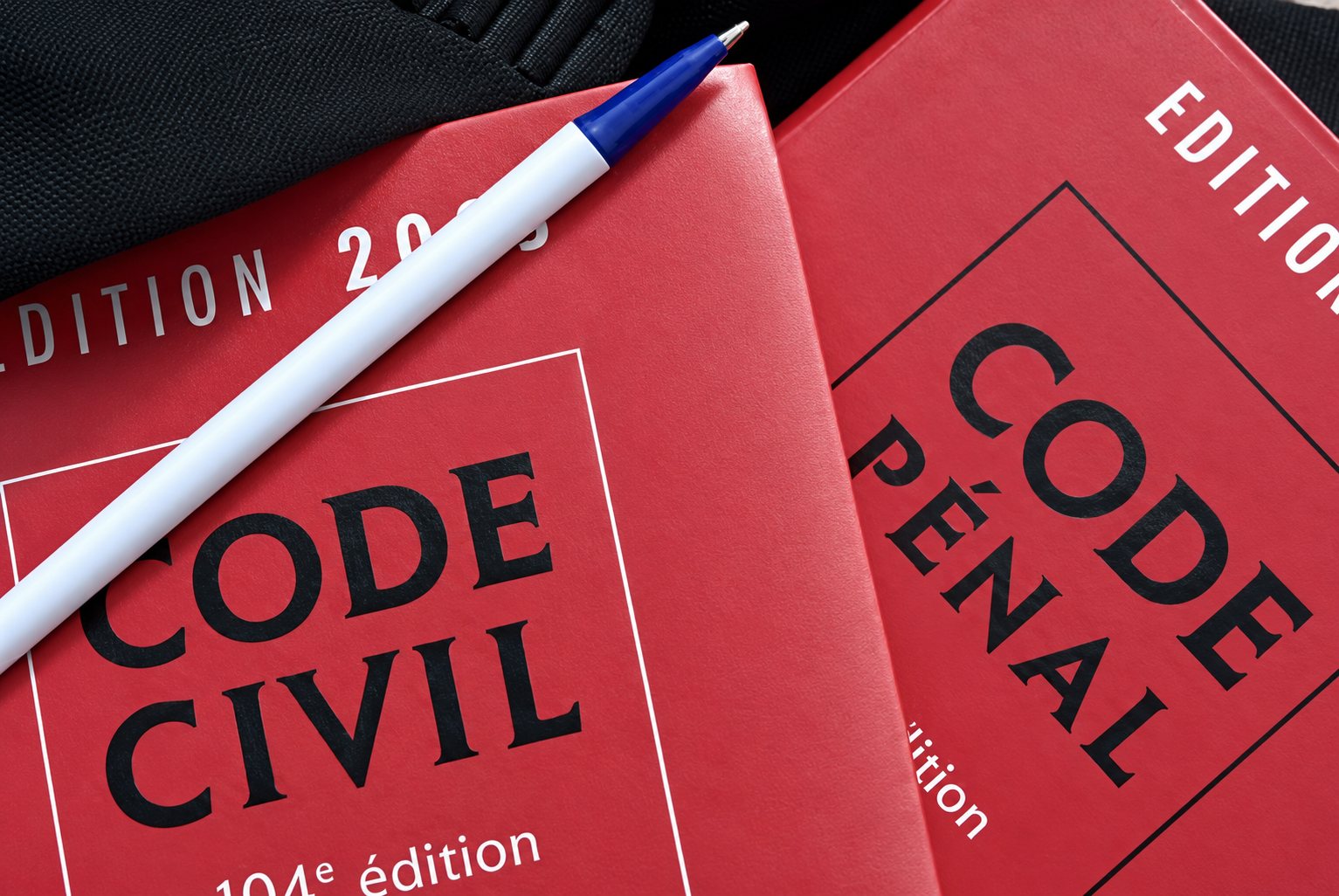 Deux ouvrages juridiques rouges, le Code civil et le Code pénal, posés l'un à côté de l'autre avec un stylo à bille blanc et bleu en travers.